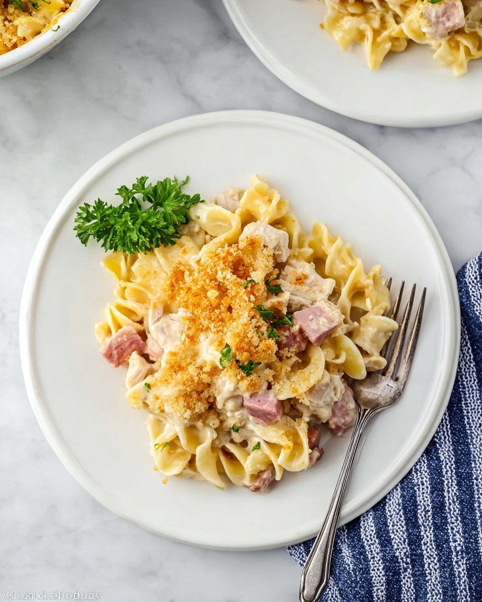 A white plate sits on a white marbled surface with a serving of baked pasta on it. The pasta has about three layers: the bottom layer is curly egg noodles with a pale yellow color, the middle layer has light pink chunks of ham and white pieces of cooked chicken, and the top layer is a golden brown breadcrumb crust with melted cheese visible underneath. A small green parsley sprig is placed on the left side of the plate, adding a fresh contrast. A silver fork rests on the right edge of the plate, partially inserted into the pasta. Part of another white plate with a similar pasta dish is visible in the upper right corner. A blue and white striped cloth napkin is seen to the bottom right on the marble surface. photo taken with an iphone --ar 4:5 --v 7