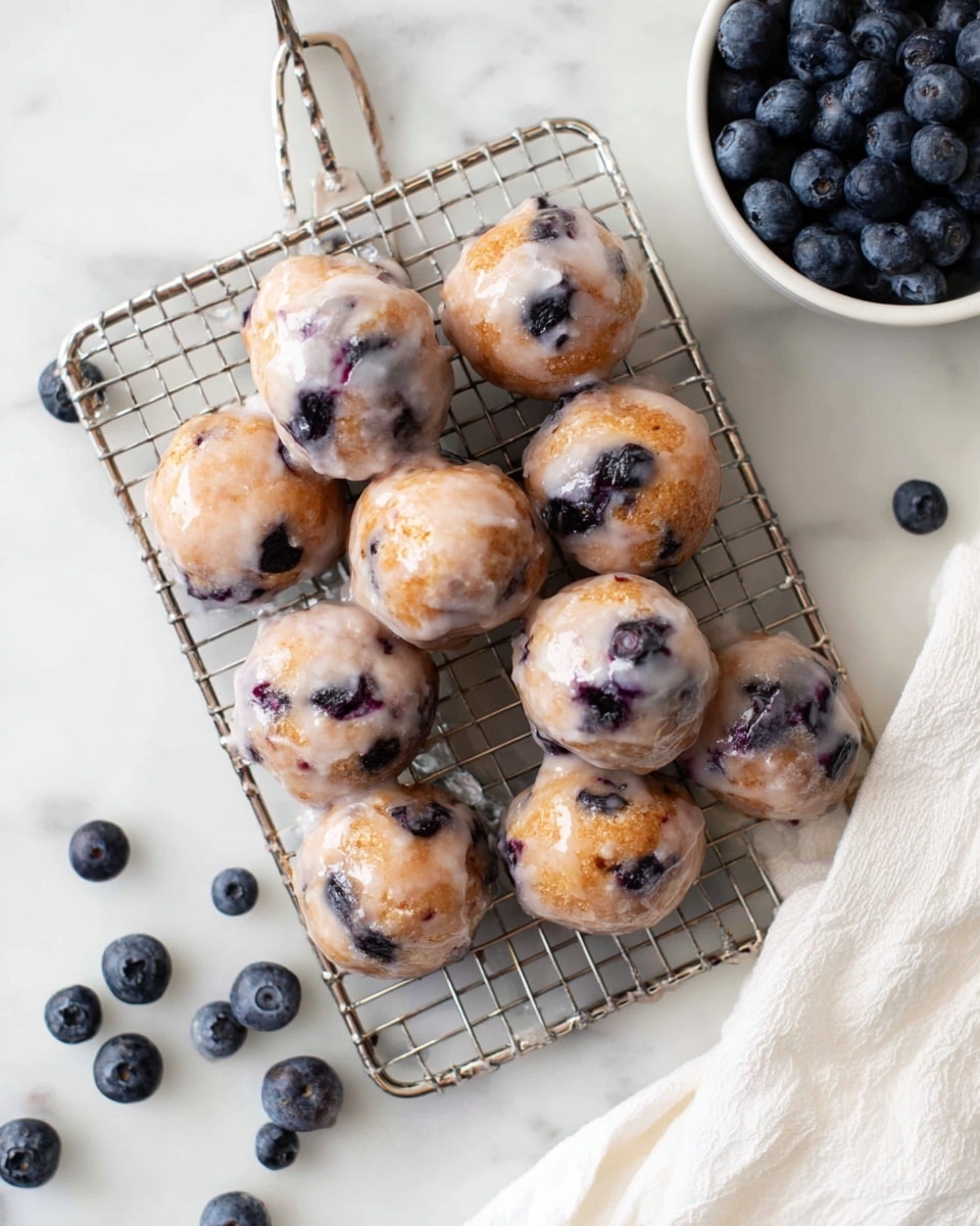 A metal wire rack holds a single layer of round blueberry doughnut holes, each covered in a shiny light glaze that gives a slightly wet texture. The doughnut holes have a pale golden color with dark purple blueberries showing through the soft dough. The wire rack is placed on a white marbled surface scattered lightly with fresh blueberries, and a white cloth is seen on the right side of the image. In the top right corner, there is a white bowl filled with more fresh blueberries. photo taken with an iphone --ar 4:5 --v 7