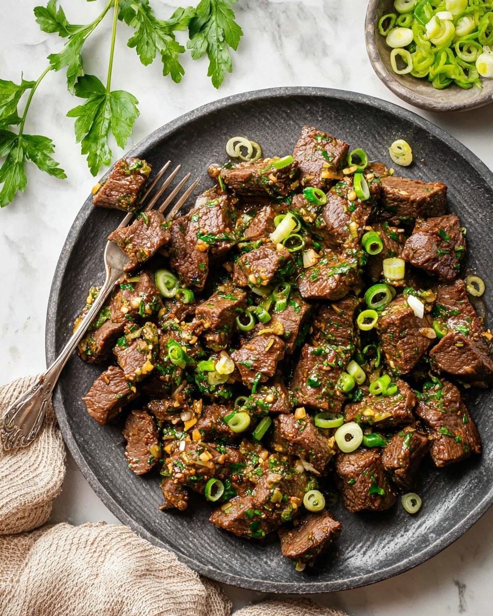 A dark grey plate holds many pieces of cooked beef cut into small chunks with a slightly shiny texture, mixed with visible green herbs and small bits of garlic, all coated in a light sauce. Thin slices of light green onion rings are scattered on top, adding a fresh color contrast. On the left side of the plate, a silver fork rests with a few pieces of beef near it. There are green parsley leaves scattered on the white marbled surface around the plate, and a small bowl with more sliced green onions is partly visible at the top right corner. A beige cloth is placed at the bottom left corner. photo taken with an iphone --ar 4:5 --v 7