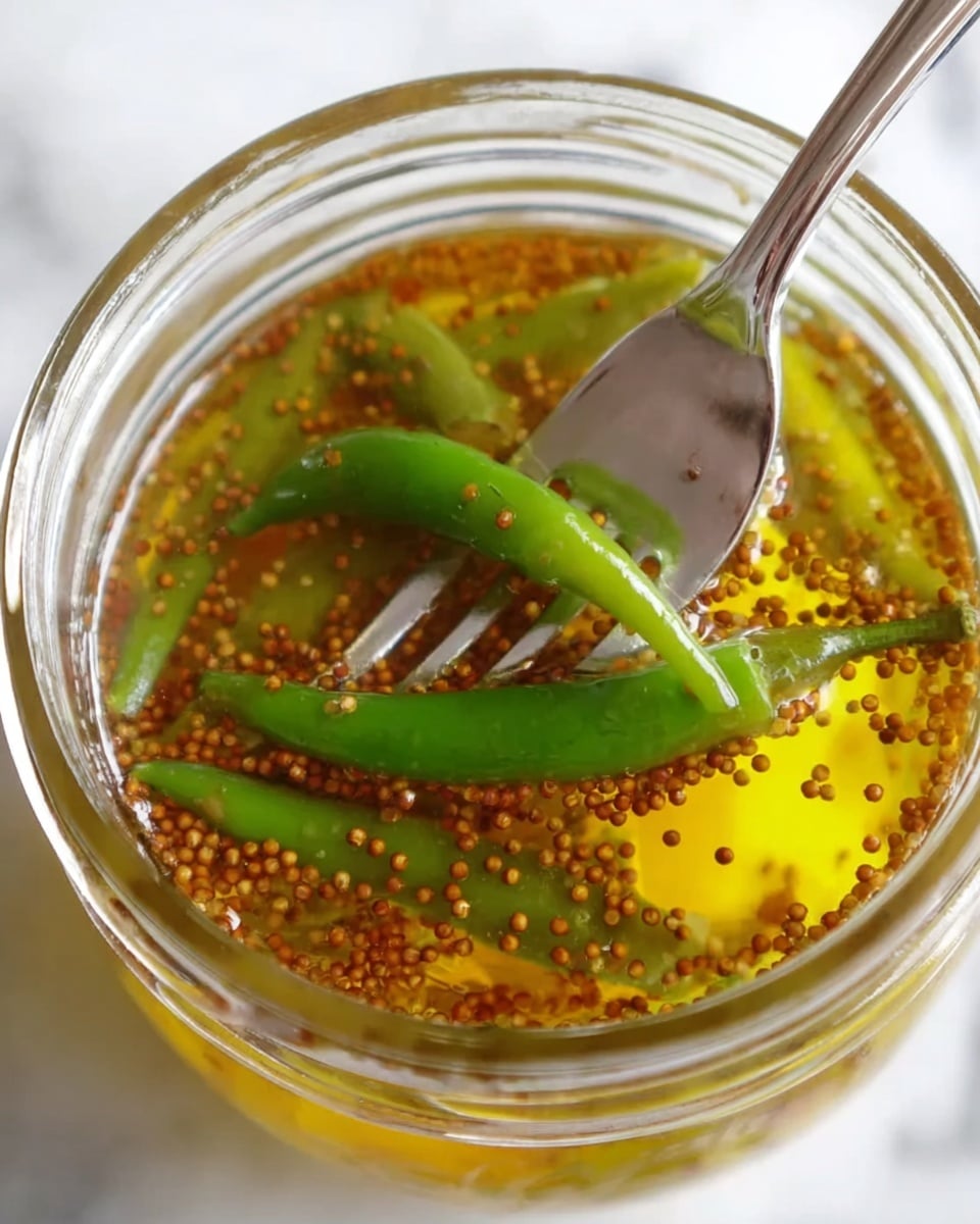 A close-up image showing a glass jar filled with golden-yellow liquid mixed with small brown mustard seeds. Inside the jar, there are several green chili peppers floating in the liquid, and a silver fork is holding one green chili pepper above the jar. The background and surface are white marbled texture. The photo taken with an iphone --ar 4:5 --v 7
