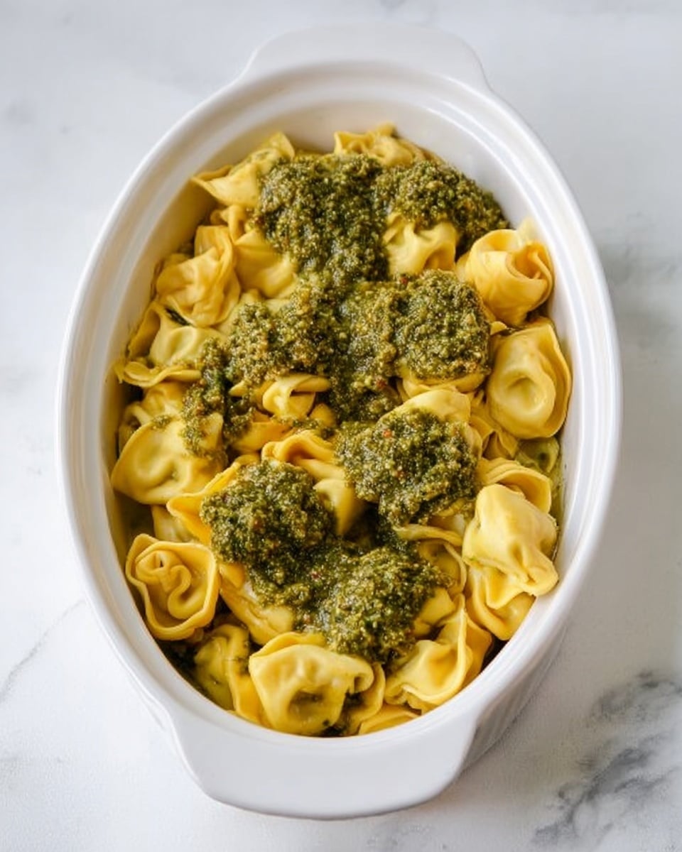 The image shows a white oval dish filled with cooked tortellini pasta that is yellow and slightly curled. On top of the pasta, there are several dollops of green pesto sauce with a coarse texture, spread unevenly over the pasta. The dish is placed on a white marbled surface. The photo taken with an iphone --ar 4:5 --v 7