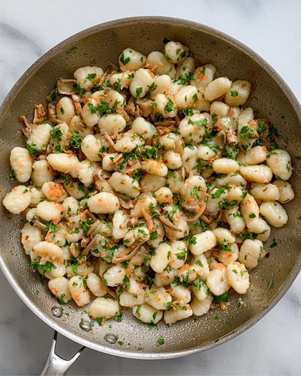 The image shows a silver frying pan filled with small white gnocchi pieces mixed evenly with small light orange pieces of cooked mushrooms and sprinkled with finely chopped green herbs. The texture of the gnocchi is soft and slightly shiny, while the mushrooms look tender and the herbs add a fresh green color on top. The pan is placed on a white marbled surface, and the colors of the food contrast nicely with the plain metal of the pan. photo taken with an iphone --ar 4:5 --v 7