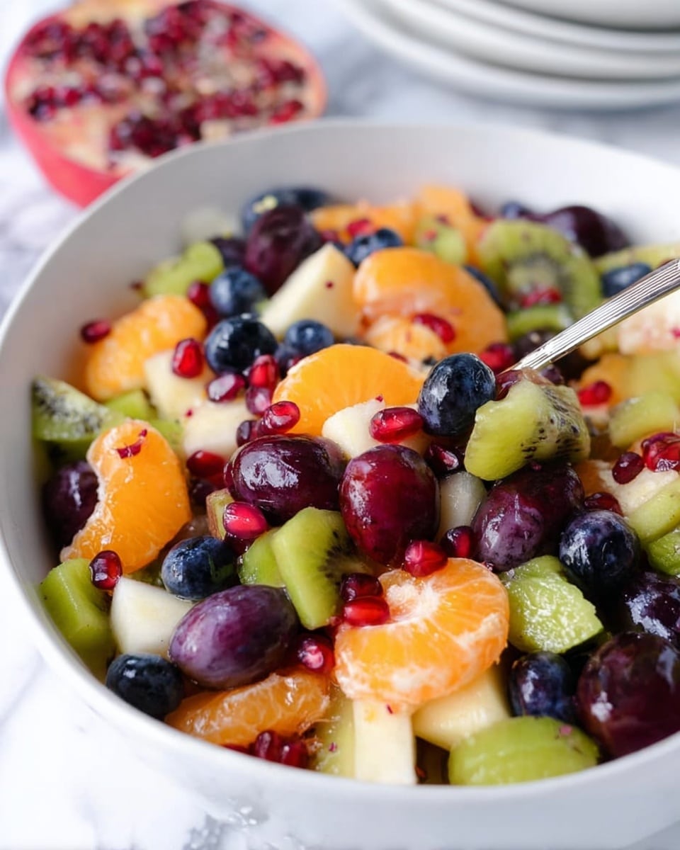A close-up of a white bowl filled with colorful fruit salad in a mix of small, bite-size pieces: bright orange mandarin slices, deep purple grapes cut in halves, green kiwifruit chunks with visible seeds, shiny dark blue blueberries, white pear pieces, and scattered bright red pomegranate seeds on top. A silver spoon is partly buried in the salad on the right side. The bowl sits on a white marbled surface with a cut pomegranate and stacked white bowls blurred in the background. Photo taken with an iphone --ar 4:5 --v 7