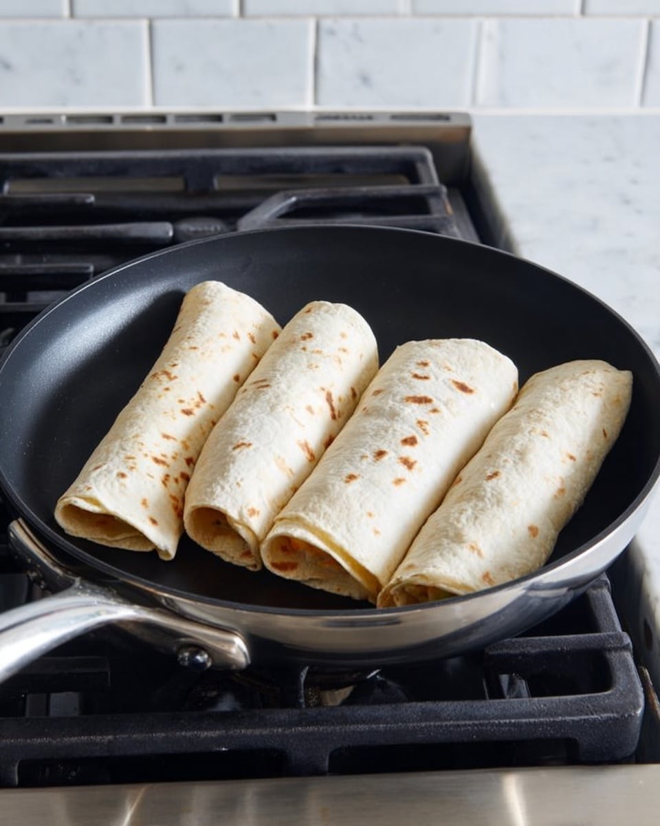 A black frying pan sits on a stove with four rolled tortillas inside it. Each tortilla is light beige with a few small brown spots, folded tightly into a rectangular shape. The frying pan is round with a shiny silver handle extending to the left. The stove has black grates, and the surface around it is white marbled texture with a tiled white backsplash in the background. The tortillas look soft and fresh, arranged in a loose square pattern around the center of the pan. Photo taken with an iphone --ar 4:5 --v 7