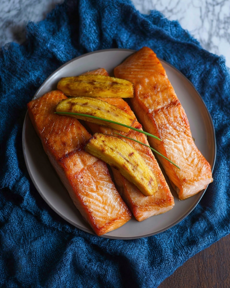 The image shows three thick orange salmon fillets arranged side by side in a row on a white plate. On top of the salmon fillets, there are three pieces of golden brown grilled plantains placed diagonally. There is a single green chive lying horizontally across the middle of the salmon fillets just below the plantains. The plate is set on a textured blue cloth, and the background has a white marbled look. photo taken with an iphone --ar 4:5 --v 7