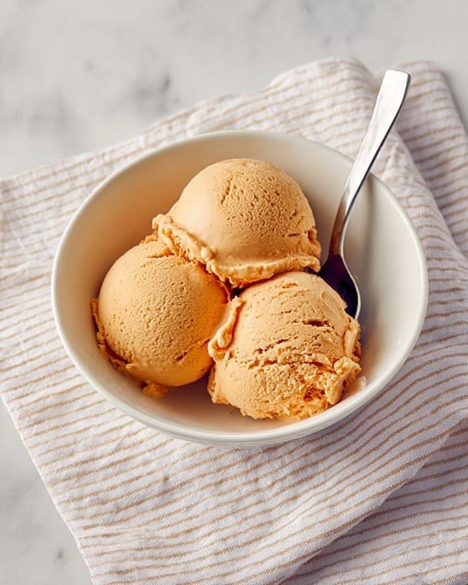 A white bowl holds three scoops of smooth, light brown ice cream. The scoops are rounded and closely packed, showing soft texture with a few small cracks. A silver spoon with a white handle rests on the edge of the bowl, pointing to the right. The bowl is placed on a white cloth with thin beige stripes, all set against a white marbled surface. photo taken with an iphone --ar 4:5 --v 7
