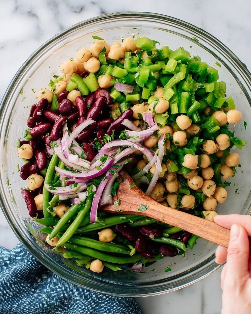 The image shows a clear glass bowl filled with a colorful bean salad. There are three main layers of ingredients: bright green chopped celery and green beans, creamy beige chickpeas, and dark red kidney beans. Thin slices of light purple onion are scattered throughout, adding a touch of soft texture. Fresh green herbs are mixed all over, giving the salad a fresh look. A woman's hand is holding a wooden spoon, stirring the salad. The bowl rests on a white marbled surface, with a blue cloth nearby. photo taken with an iphone --ar 4:5 --v 7