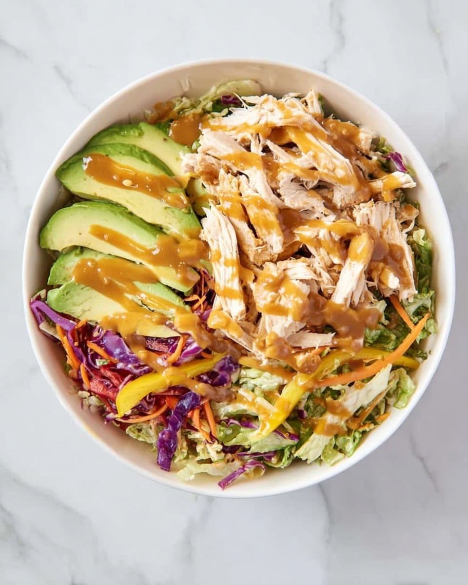 A white bowl contains a colorful salad with multiple layers: at the bottom, shredded green and purple cabbage and thin strips of carrot and yellow pepper form a leafy base. On top of this base, there are slices of light green avocado and white, shredded chicken pieces scattered evenly. The whole salad is drizzled with a golden-brown sauce that appears smooth and slightly thick. The bowl is placed on a white marbled surface, enhancing the fresh and bright look of the dish photo taken with an iphone --ar 4:5 --v 7