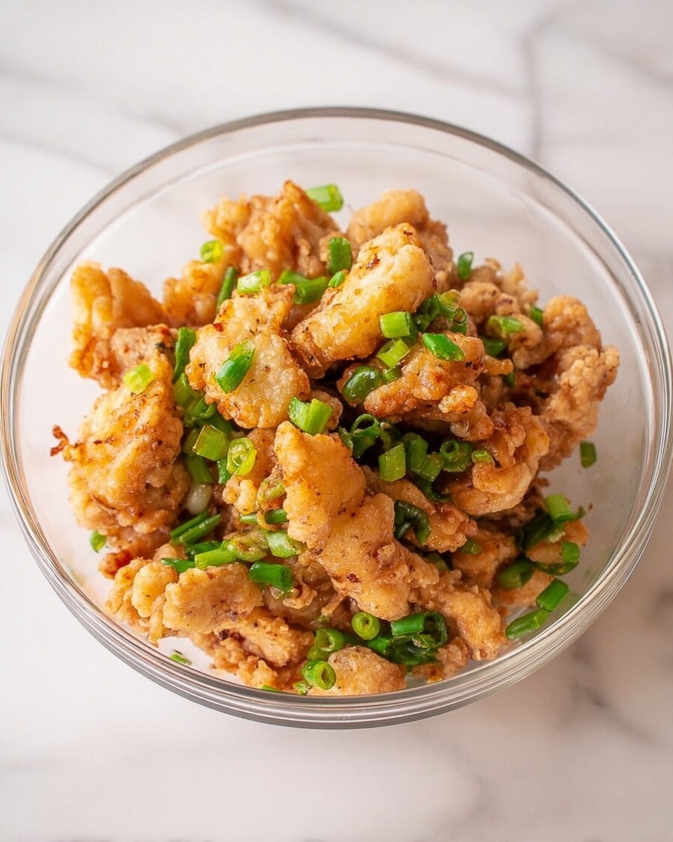 A clear glass bowl filled with about three layers of bite-sized golden fried chicken pieces mixed evenly with chopped bright green scallions. The chicken pieces have a crispy texture with some light browning, and the scallions add fresh green color scattered on top and throughout the bowl. The bowl sits on a white marbled surface, showing a clean and simple background. photo taken with an iphone --ar 4:5 --v 7