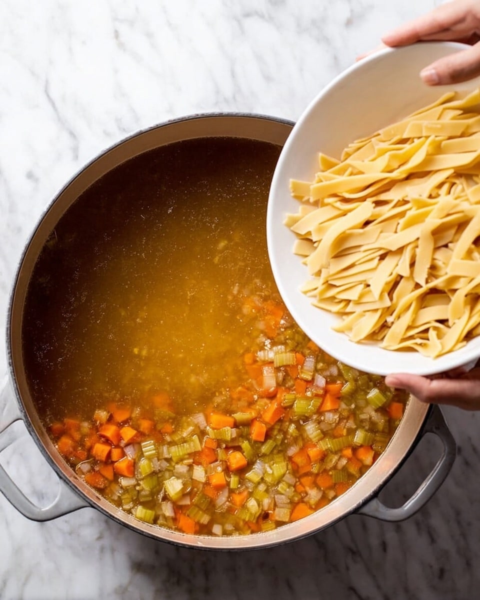 A large grey pot filled with clear golden broth that has small orange carrot cubes and green celery pieces floating inside. Above the pot, a white bowl is tilted, and a woman's hand holds it, pouring uncooked thick pale yellow pasta strips into the broth. The entire scene is set on a white marbled surface. photo taken with an iphone --ar 4:5 --v 7