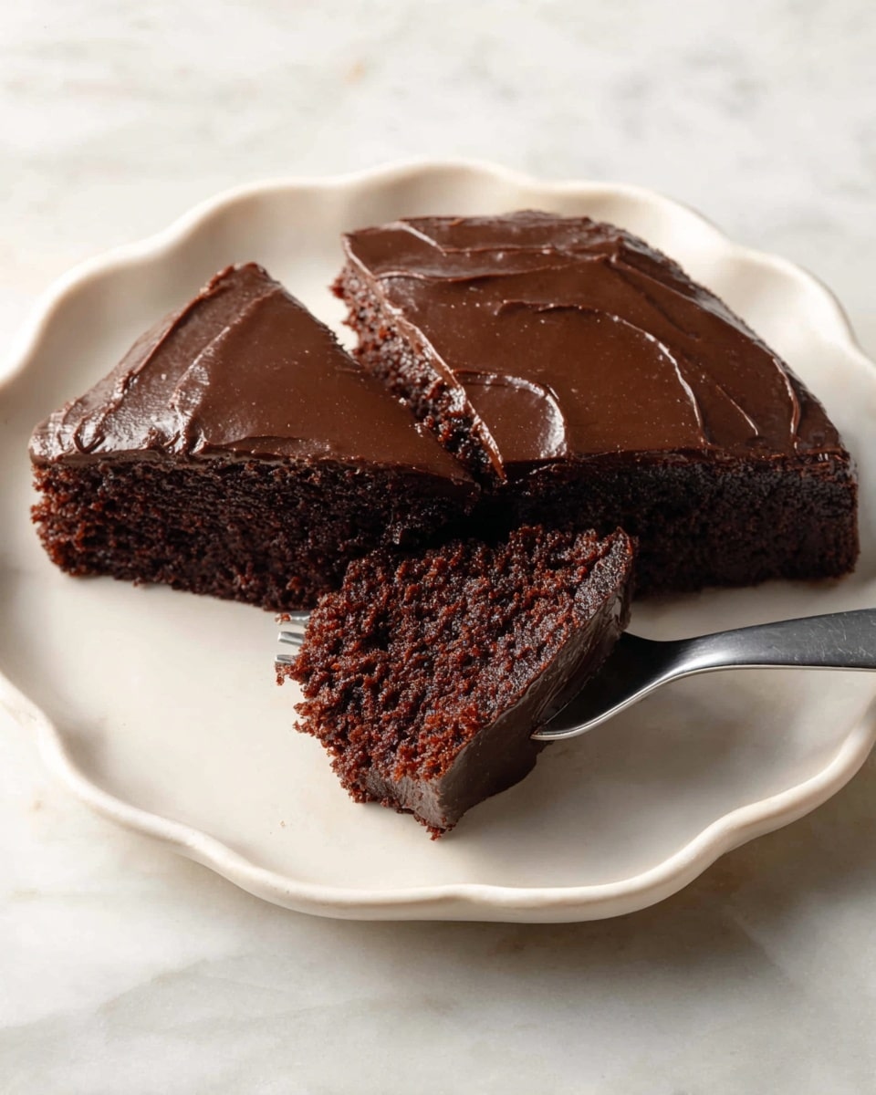 A white plate with a wavy edge holds a piece of rich, dark brown chocolate cake with thick, smooth chocolate frosting spread evenly on top. The cake is cut into two portions, one slightly angled away from the other, revealing a moist, dense interior of the same dark brown color beneath the glossy frosting layer. A metal fork is lifting a small triangle from the front portion of the cake, showing the texture of the cake inside. The plate sits on a white marbled surface. Photo taken with an iphone --ar 4:5 --v 7