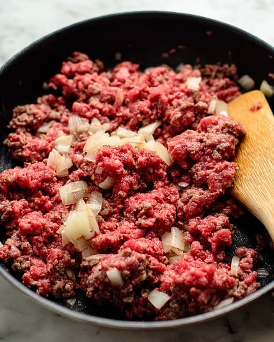 A close-up view of a black pan filled with raw and partially cooked ground meat mixed with small pale chunks of onion. The meat is unevenly cooked, showing both bright red raw sections and brown cooked parts, mixed together with the white diced onion. A wooden spatula with a light brown color rests in the pan on the right side, partially stirring the meat and onions. The pan is set on a white marbled surface. photo taken with an iphone --ar 4:5 --v 7