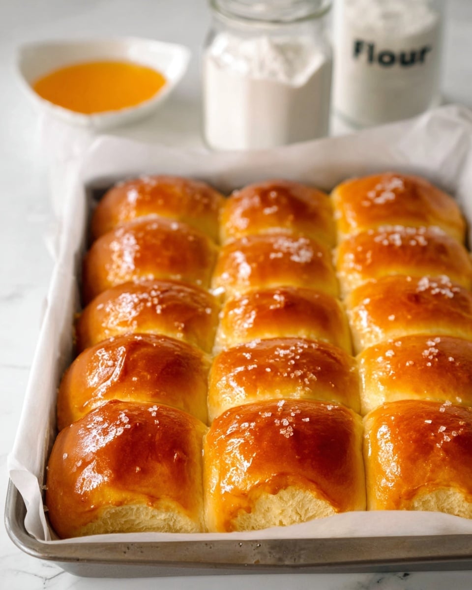 The image shows a rectangular metal baking tray lined with white parchment paper, filled with sixteen golden brown dinner rolls arranged in a 4 by 4 grid. Each roll has a shiny, smooth top with a light sprinkling of coarse salt and a soft, fluffy texture visible along the sides. In the background on the white marbled surface, there is a small white bowl filled with orange butter and a glass jar with white flour labeled
