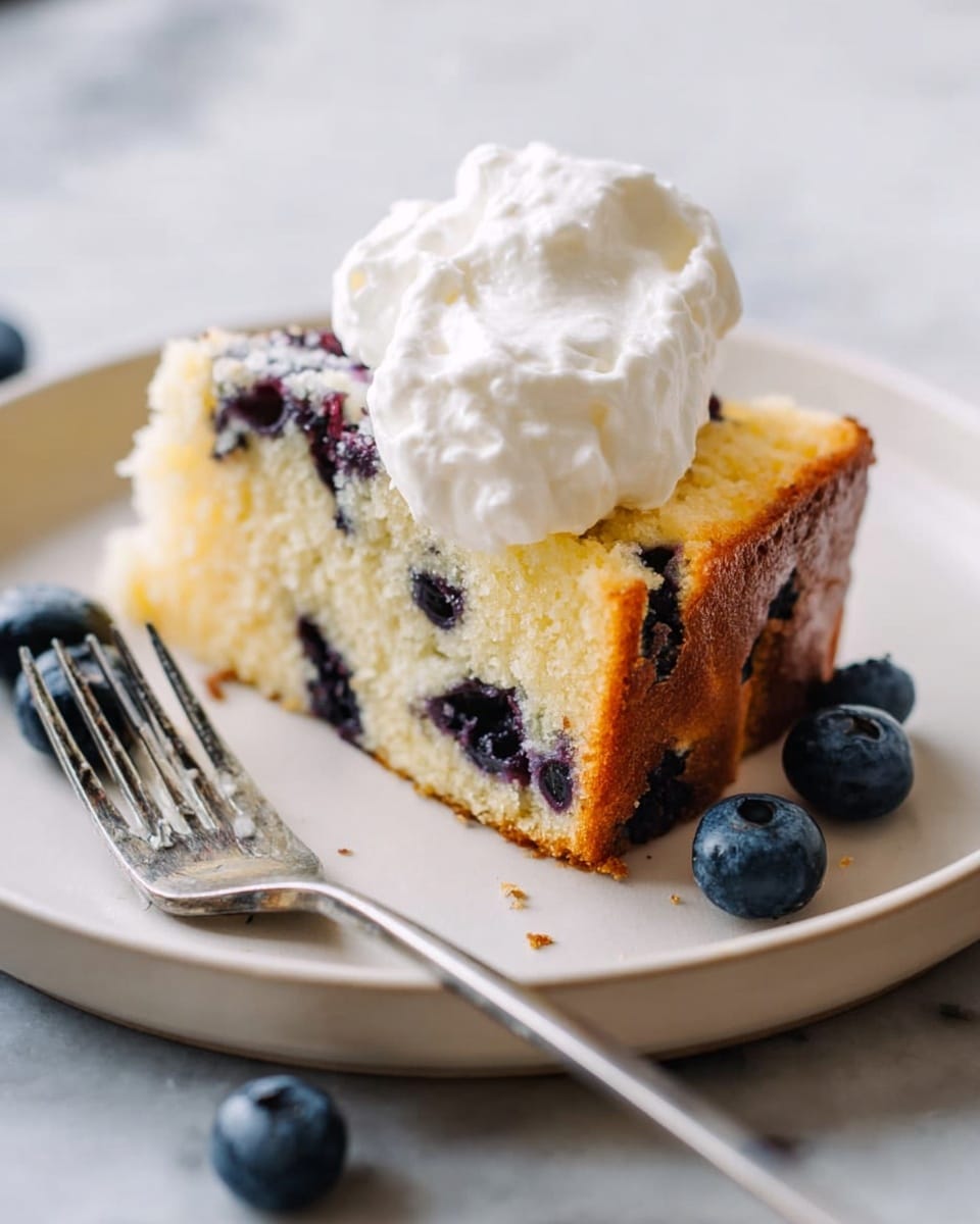 A white plate holds a slice of blueberry cake with a thick, golden brown crust on the bottom and sides. The cake is pale yellow with visible dark blueberries scattered inside. On top of the cake, there is one big dollop of white whipped cream with a fluffy texture. Around the slice, a few loose blueberries rest on the plate. A metal fork with four tines lies on the left side of the plate. The plate is placed on a white marbled surface. Photo taken with an iphone --ar 4:5 --v 7