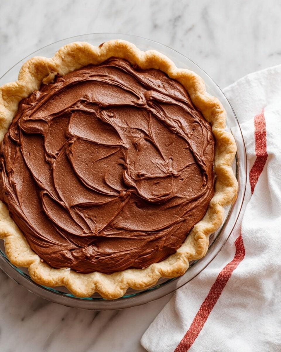 A pie with one layer of smooth, thick, dark brown chocolate filling spread in a swirling pattern on top of a light golden, flaky crust with a crimped edge, all placed in a clear glass pie dish. The pie sits on a white marbled surface next to a white cloth with two red stripes. Photo taken with an iphone --ar 4:5 --v 7