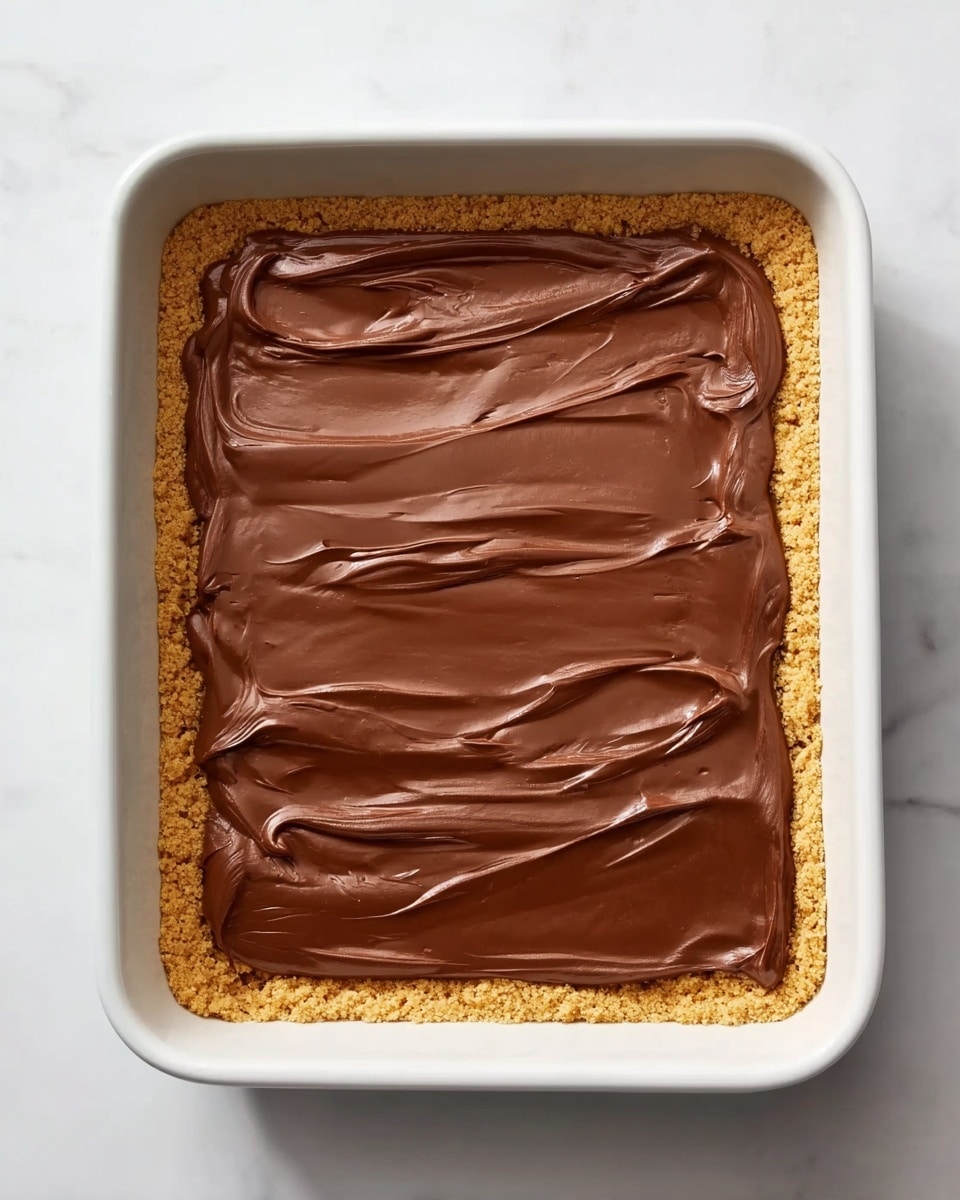 The image shows a white rectangular baking dish with a two-layer dessert inside. The bottom layer is a crumbly golden crust that evenly lines the base and edges of the dish. On top of this crust is a thick, smooth chocolate layer with a glossy texture spread carefully across, with visible swirls and slight ridges made by a spreading utensil. The dish is placed on a white marbled surface, giving a clean and bright background. photo taken with an iphone --ar 4:5 --v 7