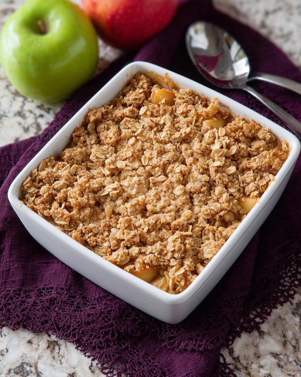 The image shows a white square baking dish filled with a crumbly apple crisp. The dish has a thick, golden-brown topping made of oats and small crumb clusters, with visible small chunks of apple peeking through. The texture of the crumble looks crunchy and slightly rough with a mix of light tan and golden colors. The dish is placed on a dark purple cloth with a lace edge, and there are two apples in the background—one red and one green—on a white marbled surface. A metal spoon lies next to the dish on the cloth. photo taken with an iphone --ar 4:5 --v 7