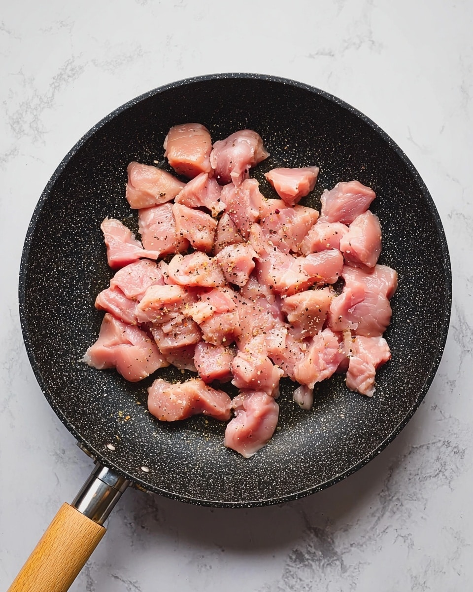 A black, speckled frying pan filled with small, raw, pink chicken pieces, lightly seasoned with black pepper, sits on a white marbled surface. The chicken pieces are spread evenly, showing a smooth but slightly glossy texture, and the pan has a wooden handle. Photo taken with an iphone --ar 4:5 --v 7