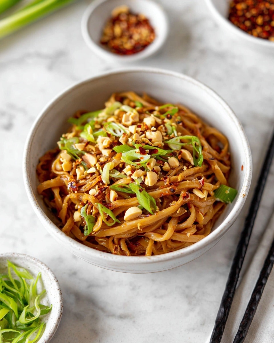 A white bowl filled with a single layer of thick, brown noodles coated in sauce, topped with scattered pieces of chopped light brown peanuts and bright green sliced scallions, along with a sprinkle of red chili flakes adding small spots of red color. The bowl sits on a white marbled surface with a pair of black chopsticks resting on the side. In the background, there are small white bowls with green scallions and red chili flakes. Photo taken with an iphone --ar 4:5 --v 7