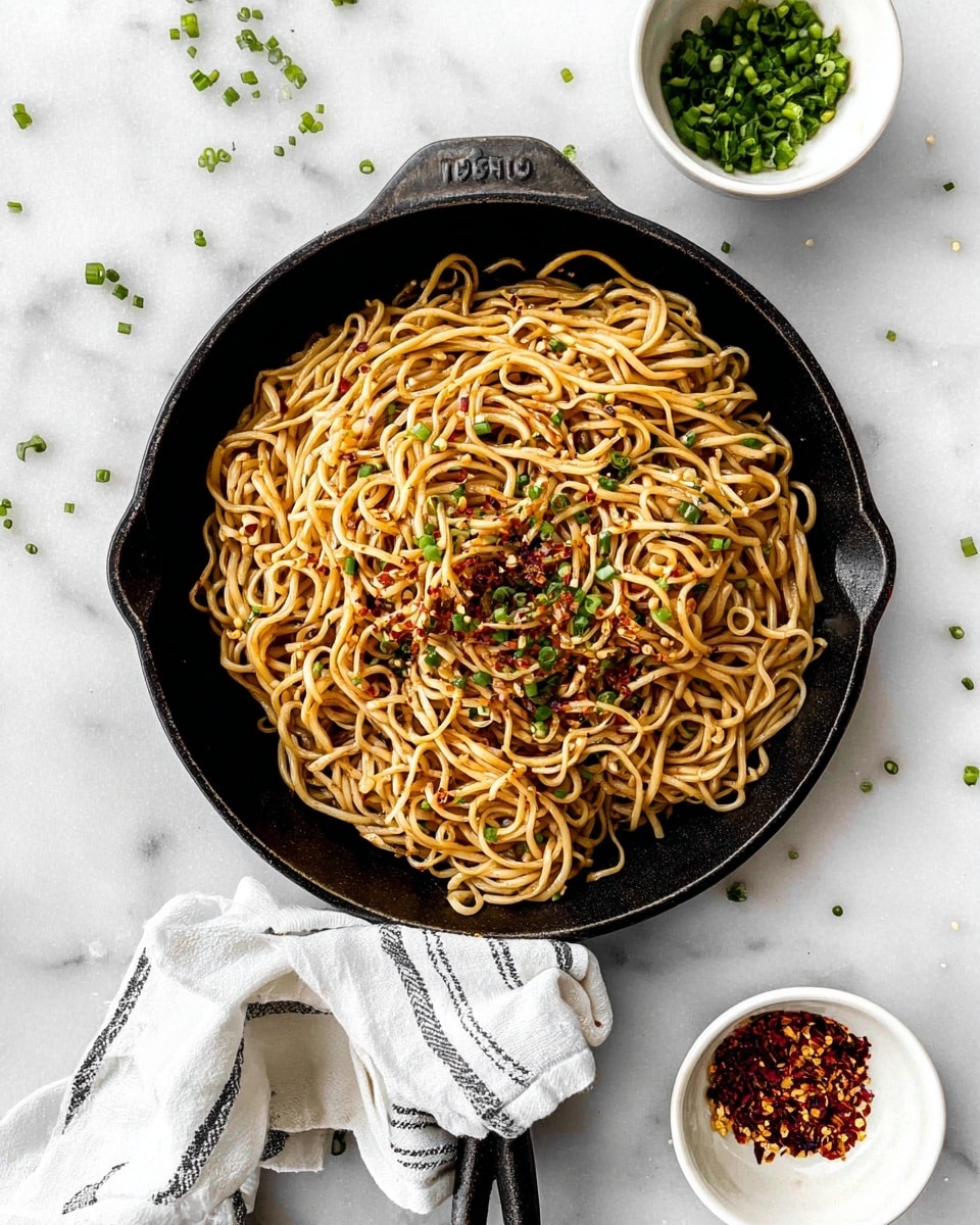 A black cast iron pan holds a single layer of cooked noodles mixed with a light brown sauce, giving them a glossy look. The noodles are garnished with small green chive pieces and a sprinkling of red chili flakes scattered mainly in the center and a few on the sides. The pan handle is wrapped in a white cloth with thin black stripes, resting on a white marbled surface. Nearby, two small white bowls sit on the surface, one filled with chopped chives and the other with red chili flakes. Some chive pieces are scattered lightly around the pan. photo taken with an iphone --ar 4:5 --v 7