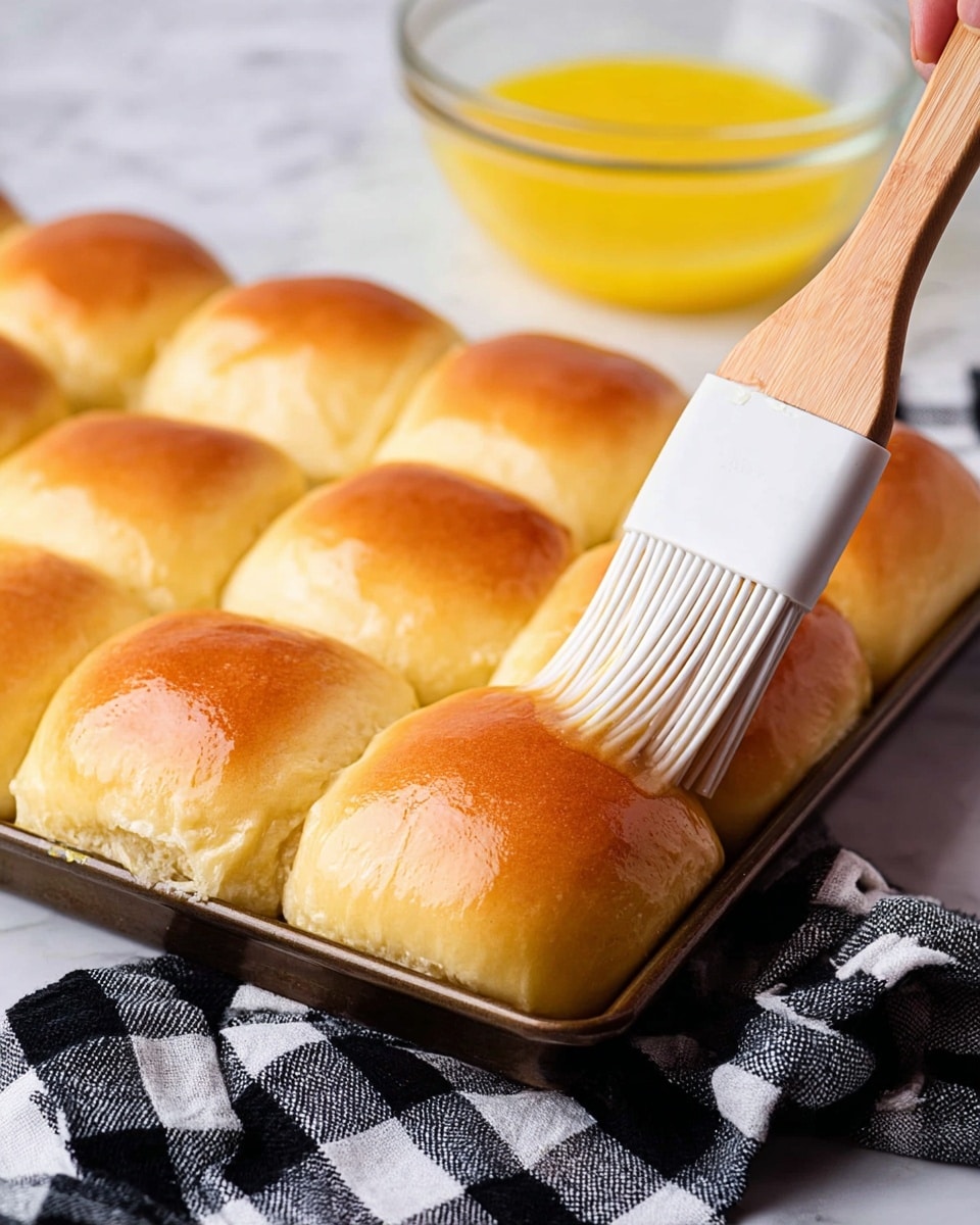 A close-up view of a baking tray filled with twelve soft, square-shaped rolls that have a shiny golden-brown top layer and a light, fluffy texture. A woman's hand brush with white silicone bristles and a wooden handle is gently applying a glaze to one of the freshly baked rolls. In the background, there is a clear glass bowl containing a yellow liquid, likely melted butter, and a black-and-white checkered cloth resting next to the tray. The scene is set on a white marbled texture surface. Photo taken with an iphone --ar 4:5 --v 7