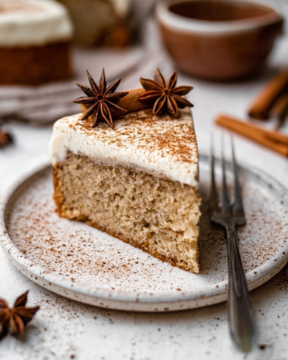 A single slice of light brown cake with a soft, fluffy texture sits on a white speckled plate. The cake has one visible layer topped with a thick, smooth white frosting layer that is dusted lightly with brown cinnamon powder. On top of the frosting, there is a decoration made of two star anise pods and a cinnamon stick placed side by side. A silver fork rests on the right side of the plate. The scene is set on a white marbled surface with blurred brown bowls and star anise in the background. Photo taken with an iphone --ar 4:5 --v 7