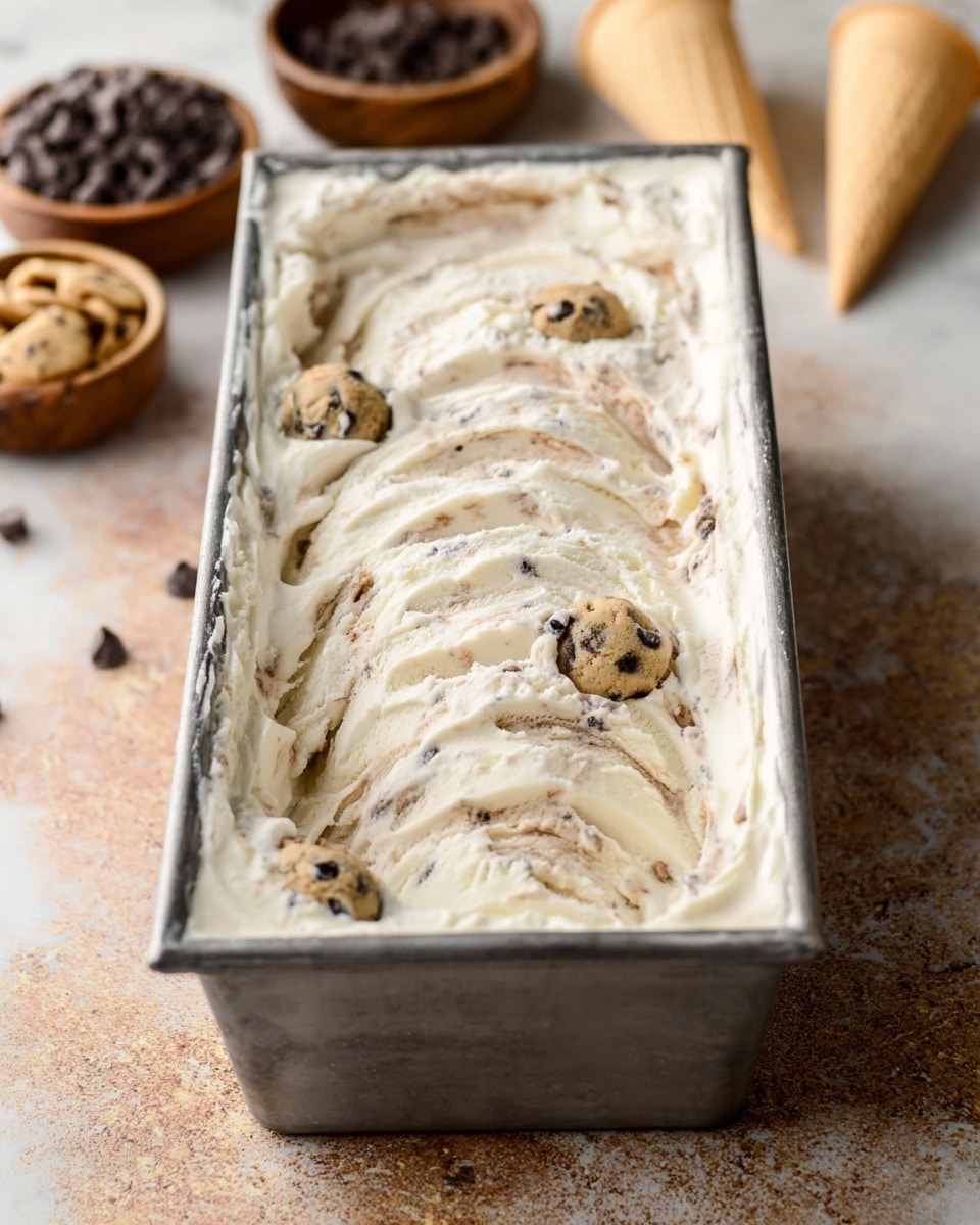 A rectangular metal container filled with creamy white ice cream that has light brown swirls mixed throughout. Visible inside the ice cream are several small round cookie dough pieces with dark chocolate chips. The container is set on a light brown textured surface, with blurred bowls of dark chocolate chips and a white waffle cone slightly visible in the background, all placed on a white marbled texture. photo taken with an iphone --ar 4:5 --v 7
