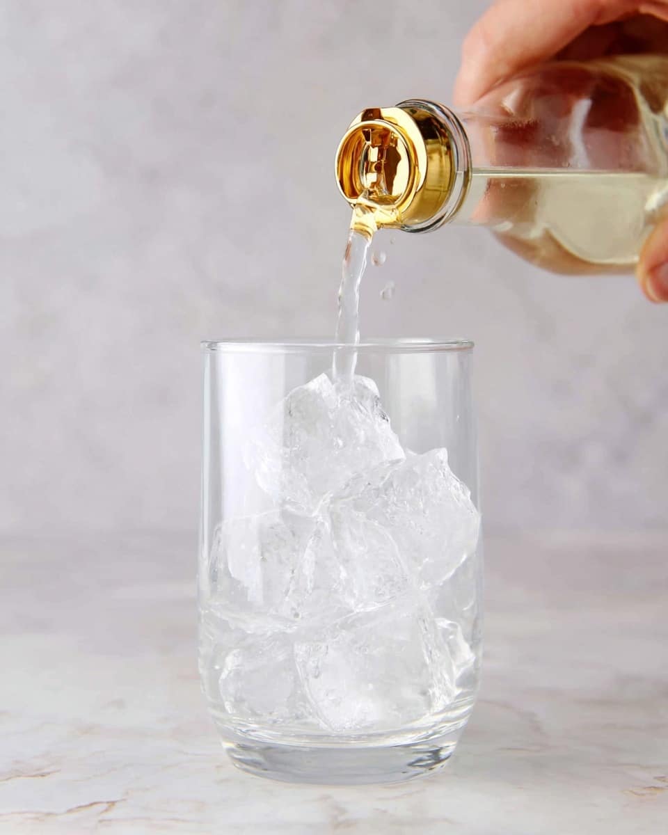 A clear glass filled with large ice cubes is shown with a light-colored liquid being poured from a bottle above it. The bottle has a gold cap with a flip-top lid open, and the liquid streams gently into the glass, creating small splashes on the ice. In the upper left corner, a woman's hand holds the bottle firmly. The background and surface are a white marbled texture, making the glass and liquid stand out clearly. photo taken with an iphone --ar 4:5 --v 7