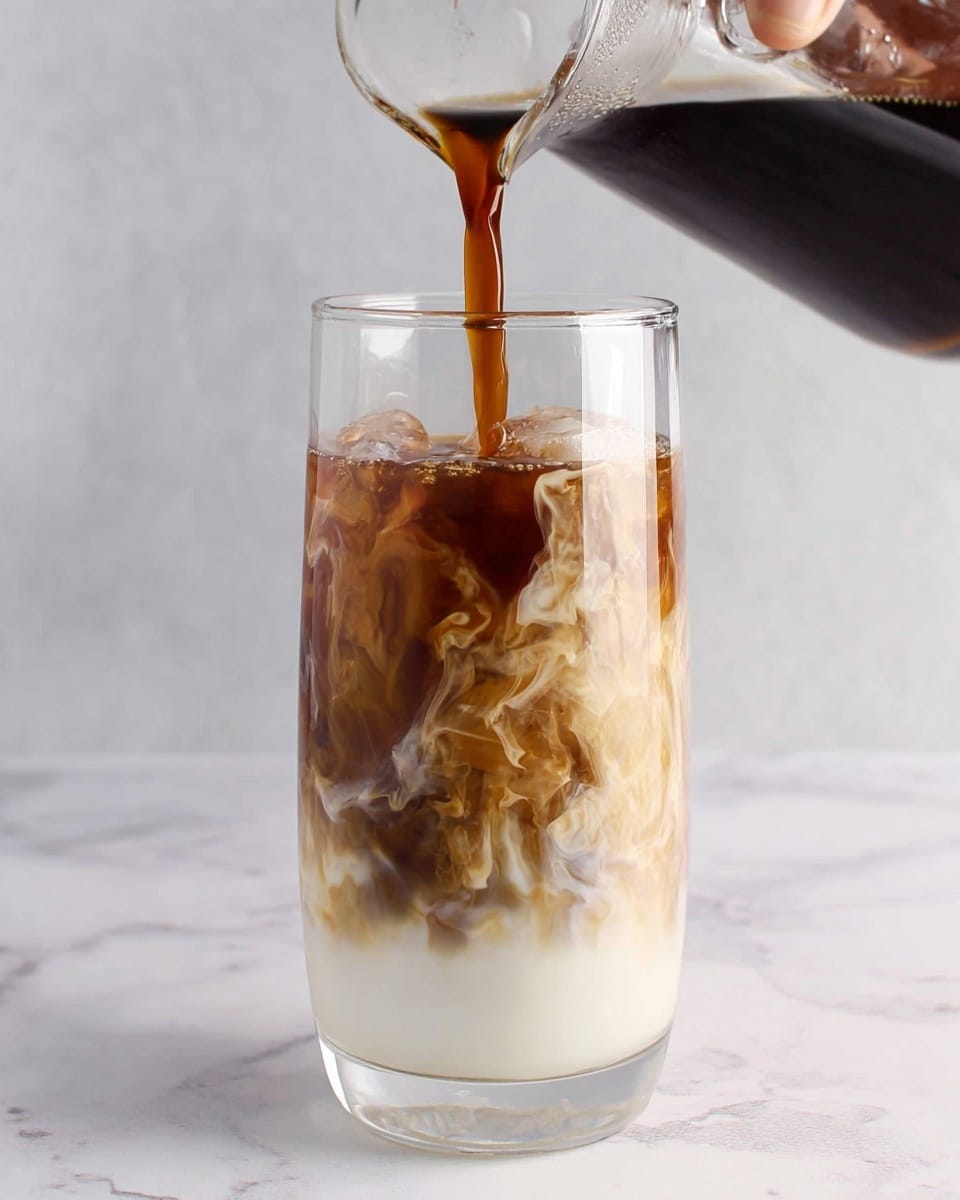 A clear glass filled with three visible layers of a drink is shown, with a woman's hand pouring dark brown coffee from a clear glass carafe into the glass. The bottom layer is white milk, smooth and opaque, taking up about half the glass. Above it, swirling patterns of light brown and white milk mix together in the middle layer, creating a cloud-like effect. The top layer is the dark brown coffee, which is being poured and mixing with the milk below. The background and surface are white marbled texture. Photo taken with an iphone --ar 4:5 --v 7