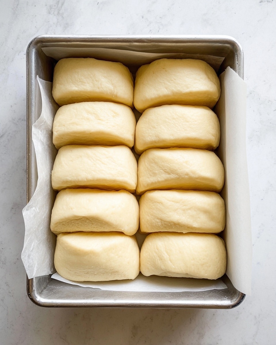 The image shows a metal baking pan lined with white parchment paper, filled with three rows of uncooked dough pieces. Each row has six soft, pale yellow dough pieces that are thick and rectangular with rounded edges, placed closely next to each other in neat lines. The dough has a smooth and slightly shiny surface, and the pan is set on a white marbled texture background. The photo taken with an iphone --ar 4:5 --v 7