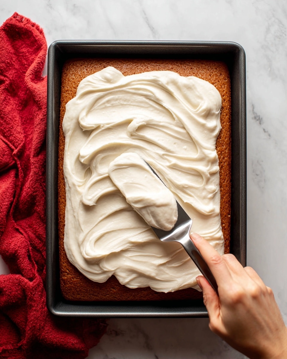 A large rectangular cake with a smooth golden-brown top sits inside a black metal baking pan. On top of the cake, there is a thick layer of white cream being spread in broad, smooth swirls by a woman's hand holding a silver spatula. The background surface is a white marbled texture, with a red cloth partially visible on the left side. The cream contrasts sharply with the warm brown cake, creating a fresh and inviting look. photo taken with an iphone --ar 4:5 --v 7