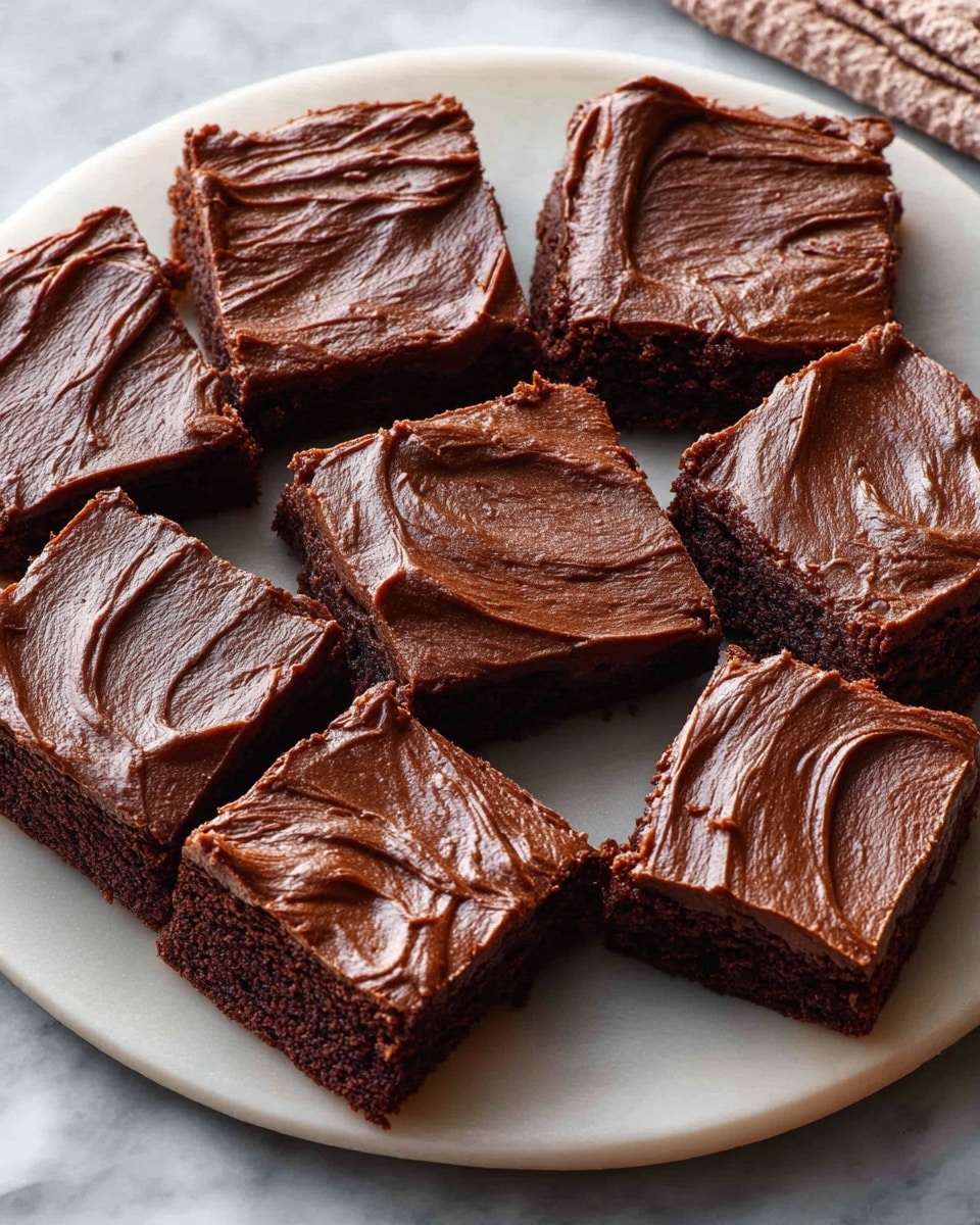 A white plate holds nine square chocolate brownies arranged closely together, with one square slightly pulled away at the front. Each brownie has a smooth, thick layer of dark chocolate frosting on top showing creamy swirls and ridges. The brownies themselves are dense and dark brown, contrasting with the glossy frosting. The plate sits on a white marbled surface, adding a clean look. Photo taken with an iphone --ar 4:5 --v 7