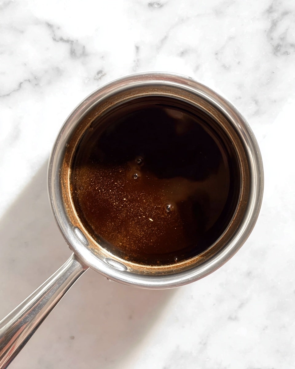 A small silver saucepan filled with a dark brown, smooth sauce sits on a white marbled surface. The sauce has a shiny texture and looks thick but liquid. The saucepan handle extends to the left side of the image. The lighting highlights the glossy surface of the sauce and the clean edges of the pan. Photo taken with an iphone --ar 4:5 --v 7
