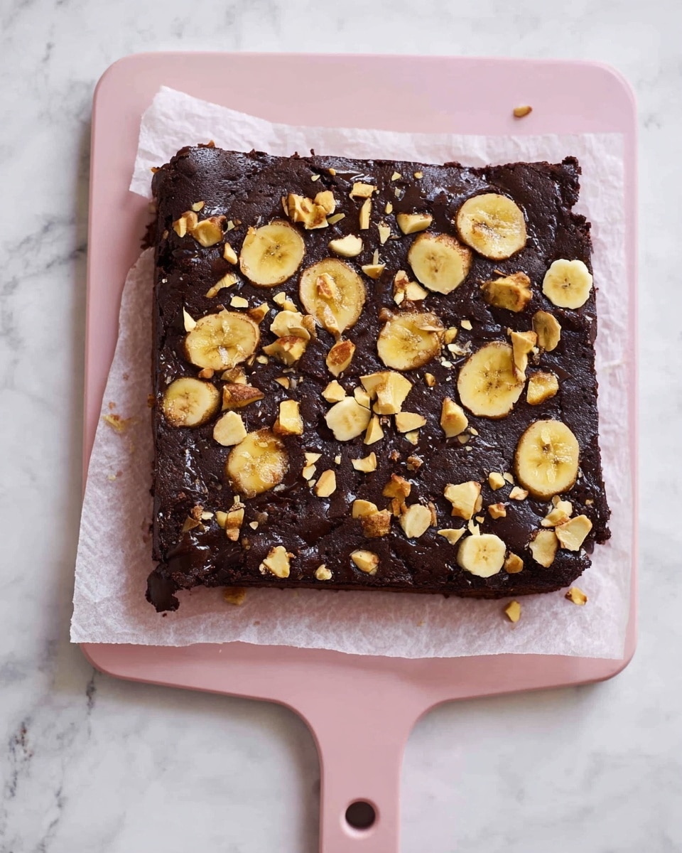 A square, dark brown chocolate brownie sits on white parchment paper placed on a light pink cutting board. The top of the brownie is sprinkled with many pieces of light yellow banana chips scattered unevenly across its rough and slightly shiny surface, adding texture and contrast. The brownie looks dense with some small cracks on the edges. The pink cutting board has a handle with a round hole near its top. The setup is on a white marbled surface. photo taken with an iphone --ar 4:5 --v 7