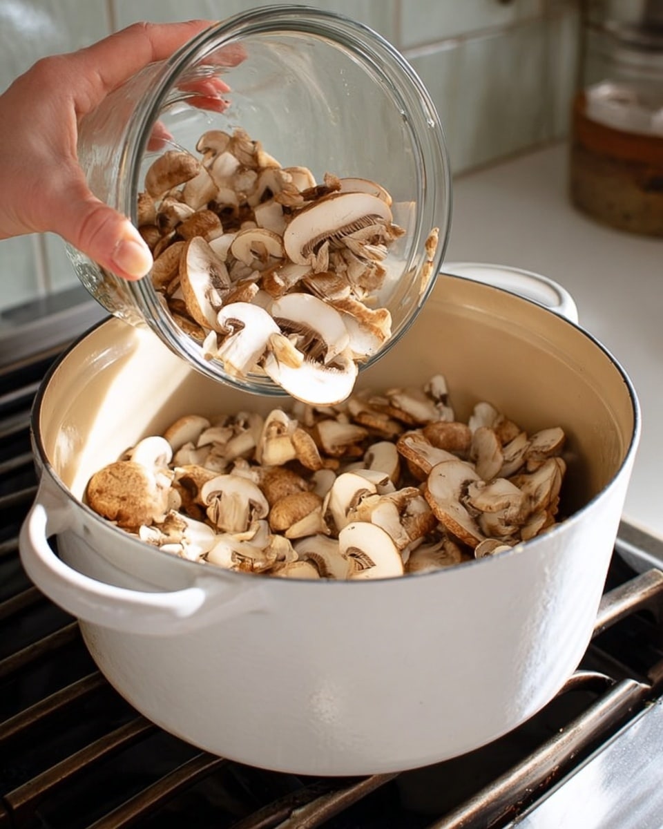 A white pot sits on a stove with a white marbled surface, filled partially with sliced brown mushrooms that have white edges and light brown caps. Above the pot, a woman's hand holds a clear glass bowl, pouring more light brown and white sliced mushrooms into the pot. The background shows the stove’s metal grates and a pale tiled wall. The image has a clean and simple cooking scene with soft natural lighting photo taken with an iphone --ar 4:5 --v 7