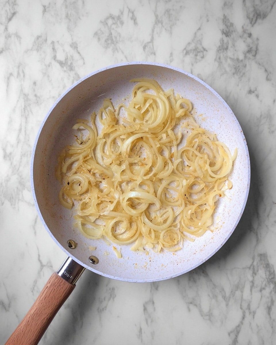 A white frying pan with a wooden handle sits on a white marbled surface, holding one layer of cooked onion slices. The onions are light golden yellow with a soft, slightly glossy texture, spread evenly across the pan’s bottom. Photo taken with an iphone --ar 4:5 --v 7