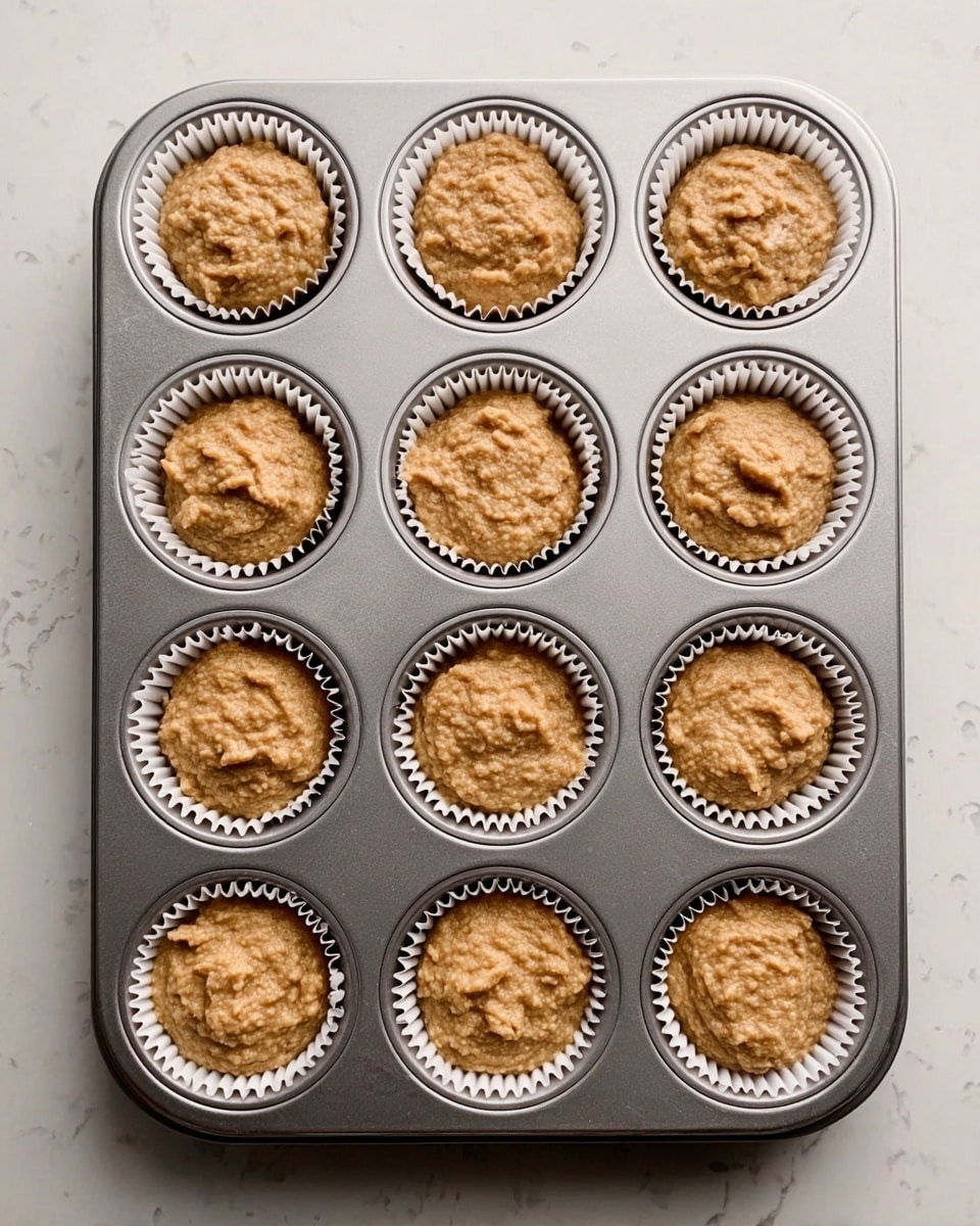 A metal muffin tray holds twelve white paper liners filled with a rough, light brown batter. Each cup is filled almost to the top with the thick, uneven-textured mixture, which looks soft and slightly grainy. The tray sits on a white marbled surface that adds a clean and bright background. There are no additional items or decorations in the image. Photo taken with an iphone --ar 4:5 --v 7