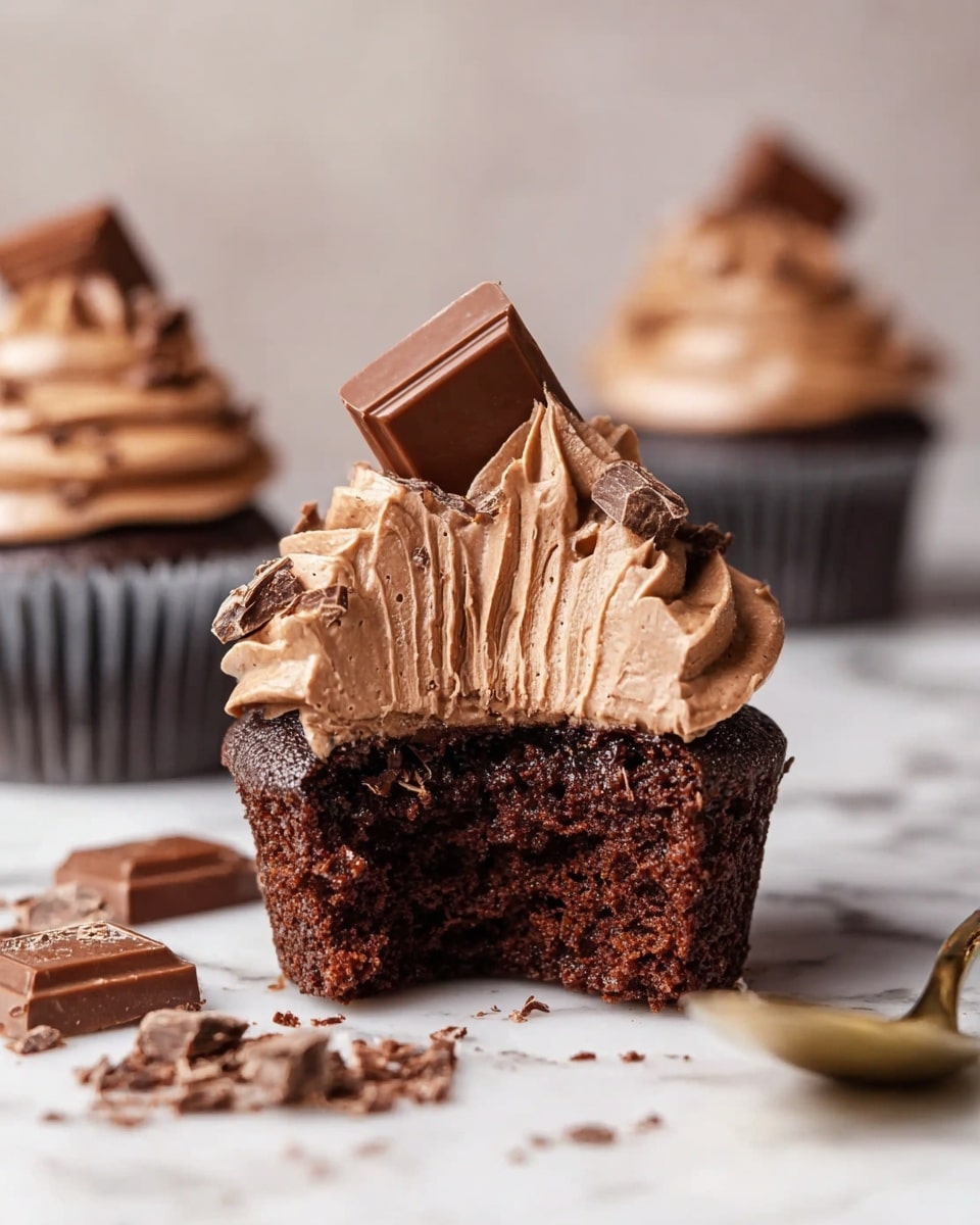 A dark brown chocolate cupcake with a moist texture is shown with a large bite taken out of it, revealing the crumb inside. On top, there is a thick swirled layer of light brown chocolate frosting with a soft, creamy texture, crowned with a small square piece of milk chocolate inserted into the frosting. Crumbs and small pieces of chocolate are scattered around the base of the cupcake on a white marbled surface. In the background, two whole cupcakes with the same frosting and chocolate square decoration sit slightly out of focus. A gold spoon with some crumbs rests near the bitten cupcake. photo taken with an iphone --ar 4:5 --v 7