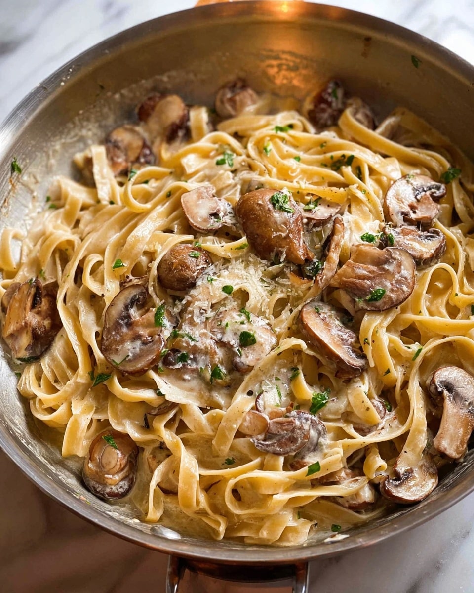 The image shows a pan of creamy pasta with mushrooms. There are two main layers: a base layer of light tan fettuccine noodles that look soft and smooth, and on top are sliced brown mushrooms, some with a creamy sauce coating. Small green herb pieces are scattered over the dish, adding color and freshness. The pan has a silver-gray metal shine and is resting on a white marbled surface. Photo taken with an iphone --ar 4:5 --v 7