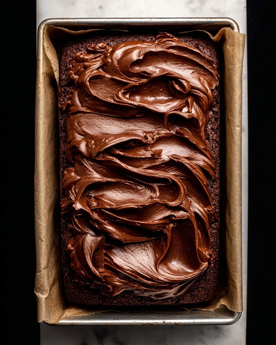 A rectangular chocolate cake sits inside a metal baking tray lined with parchment paper. It has one thick layer of dark brown chocolate cake at the bottom, topped with a generous, thick layer of shiny, smooth chocolate frosting spread in swirled ribbons with peaks and waves, giving it a rich texture. The scene is set on a white marbled surface. Photo taken with an iphone --ar 4:5 --v 7