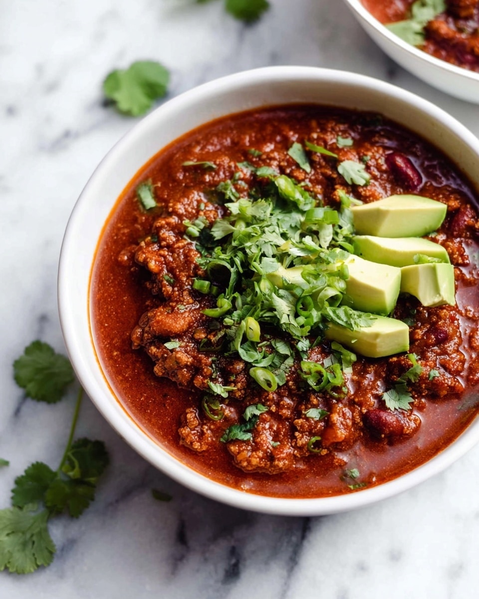 A white bowl filled with three layers: the bottom layer is thick red chili with visible chunks of meat and beans with a rich, hearty texture; the middle layer is a neat topping of sliced avocado pieces in light green; the top layer is sprinkled with fresh green herbs, mainly cilantro and green onion, adding a fresh contrast to the rich chili below. The bowl sits on a white marbled surface, with small sprigs of cilantro scattered nearby. Photo taken with an iphone --ar 4:5 --v 7