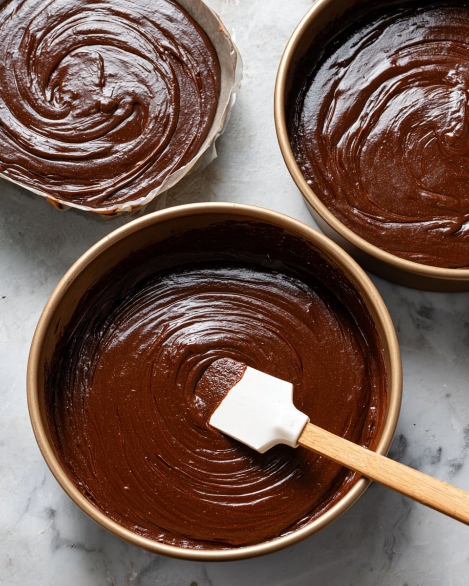 The image shows two round cake pans filled with smooth, dark chocolate batter. Each pan contains one thick layer of rich, glossy chocolate batter with visible swirls made from spreading. A wooden spatula with a white silicone head, coated in the same batter, rests inside one pan. Both pans sit on a white marbled surface with a clean and simple background. Photo taken with an iphone --ar 4:5 --v 7