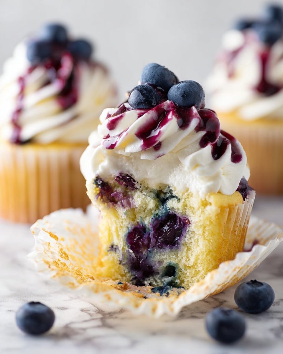 The image shows a close-up of two blueberry cupcakes on a white marbled surface. The cupcake in the front is partly unwrapped from its white paper liner, revealing a soft yellow cake with scattered whole blueberries inside. On top of this cupcake is a thick layer of white swirled cream, drizzled with dark purple blueberry sauce, and garnished with fresh blueberries. The second cupcake in the background is decorated similarly with white cream, blueberry sauce, and fresh blueberries. There are a few loose blueberries around the cupcakes. Photo taken with an iphone --ar 4:5 --v 7
