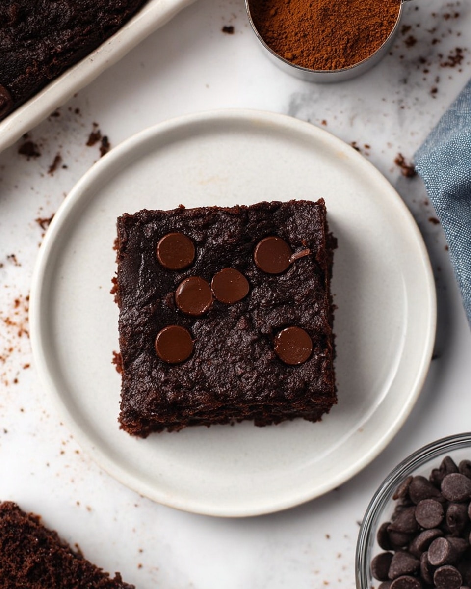 A single square piece of dark brown chocolate brownie sits centered on a white plate, showing a slightly shiny and moist surface with several large, smooth dark chocolate chips melting slightly into the top layer. The brownie looks dense and rich, with a textured edge hinting at a soft inside. Around the plate, there’s a white marbled surface with scattered chocolate chips and parts of other plates with more brownie pieces barely visible. A metal measuring cup with cocoa powder and a glass bowl filled with round chocolate chips are also partially seen. photo taken with an iphone --ar 4:5 --v 7
