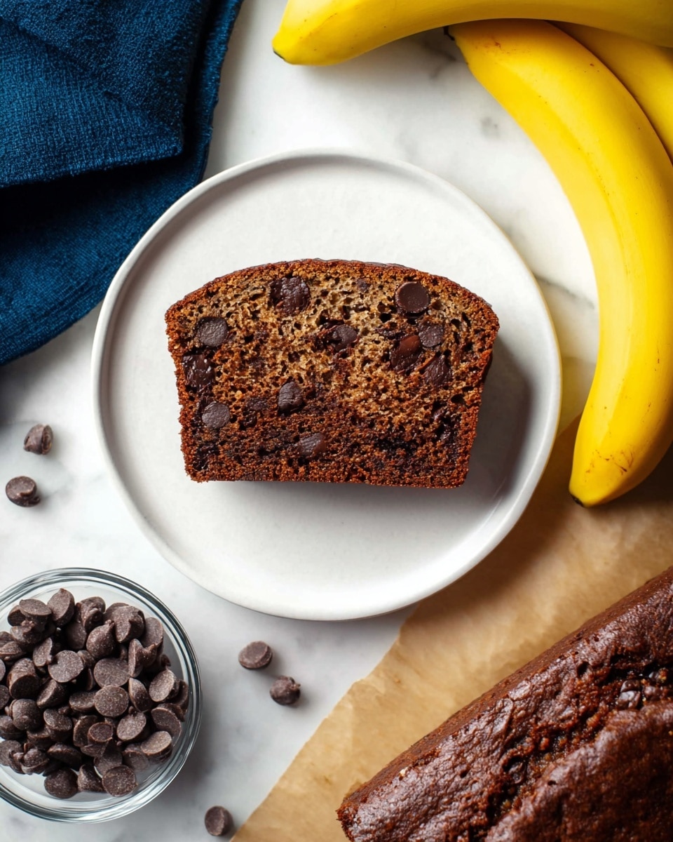 A single slice of dark chocolate banana bread with a moist texture and chocolate chips inside is centered on a white plate. To the left of the plate, there is a small clear glass bowl filled with shiny dark chocolate chips and a few chocolate chips scattered on the white marbled surface. To the right, there are two ripe yellow bananas resting against the white marbled background. The bottom right corner shows part of the remaining loaf of chocolate banana bread on brown parchment paper. A dark blue cloth is on the left side of the image. Photo taken with an iphone --ar 4:5 --v 7