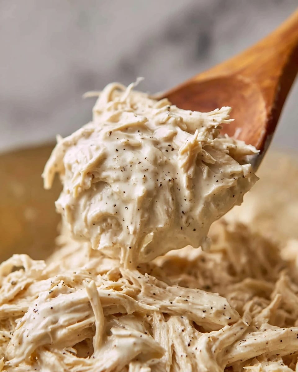 The image shows a close-up of shredded chicken mixed with a creamy white sauce, speckled with black pepper for texture and seasoning. The chicken strands are thick and soft, coated evenly by the sauce, which looks smooth and rich. A wooden spoon is lifting a generous scoop of the mixture from a larger amount in the background, showing the sauce clinging to the chicken in a slightly glossy finish. The background features a softly blurred white marbled texture, giving focus to the creamy chicken. photo taken with an iphone --ar 4:5 --v 7