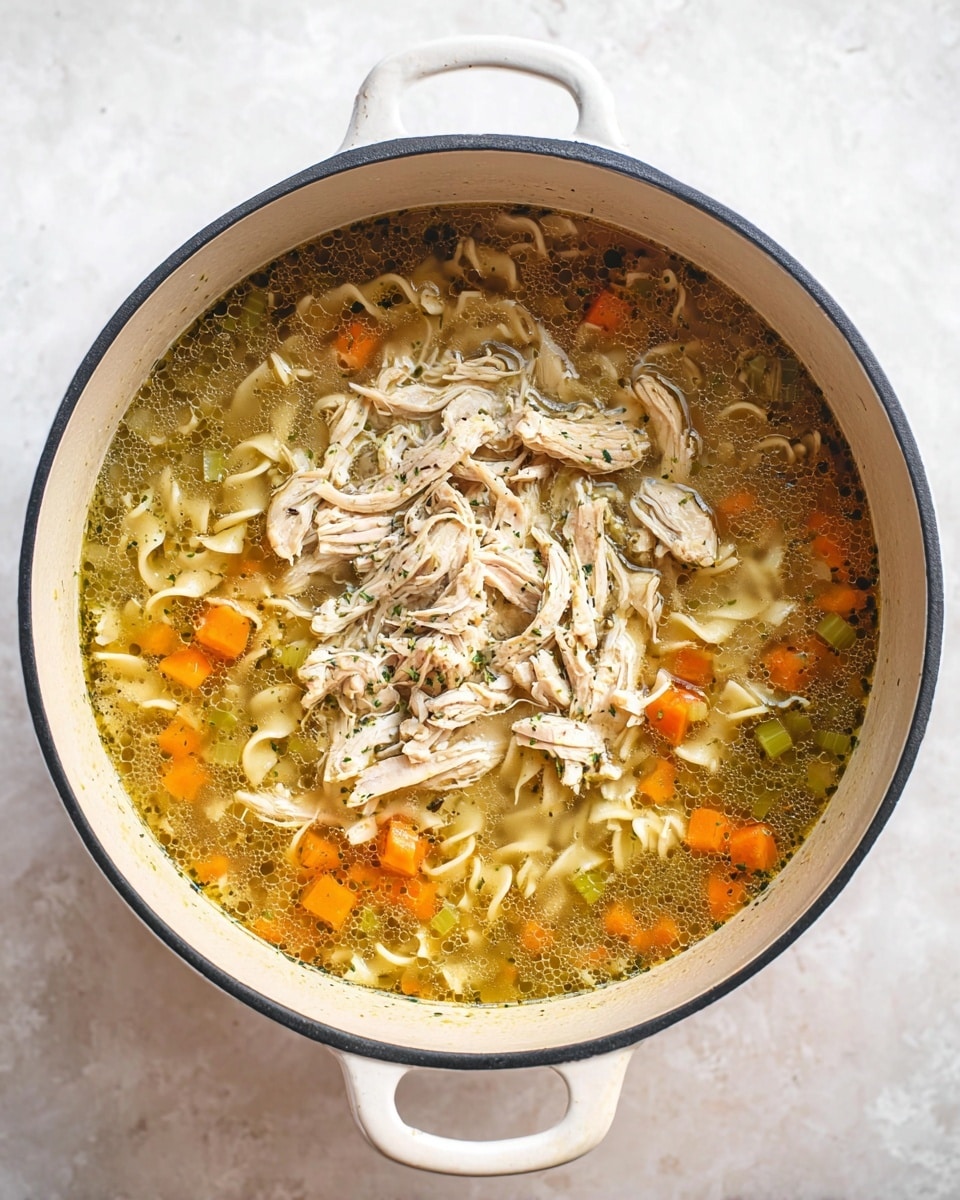 A white cast iron pot filled with clear broth soup showing three main layers: the bottom layer is a mix of soft, diced orange carrots and light green celery pieces, the middle layer has small, curly beige egg noodles scattered through the broth, and the top layer features shredded light tan chicken piled in the center. The broth has a slightly oily surface with visible herbs and small black specks of seasoning. The pot is placed on a white marbled texture surface. Photo taken with an iphone --ar 4:5 --v 7