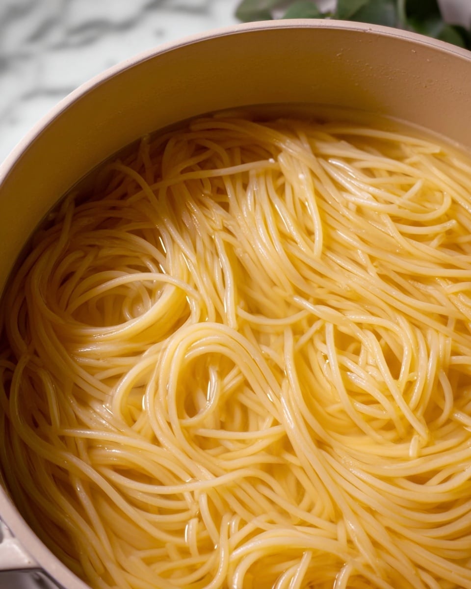 A close-up image of plain cooked spaghetti noodles submerged in clear water inside a beige pot. The noodles are light yellow with a smooth texture, loosely tangled and floating evenly throughout the pot. The background shows a white marbled surface with some green leaves in soft focus. The image has a natural, simple look with no extra toppings or sauces visible, photo taken with an iphone --ar 4:5 --v 7