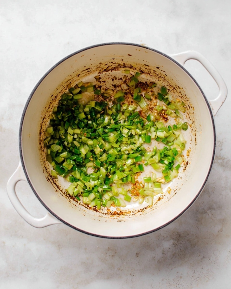 The image shows a white pot with two handles on a white marbled surface. Inside the pot, there is one layer of chopped green onions being cooked, with parts of them slightly softened and mixed with small patches of golden brown bits stuck to the bottom of the pot. The green onions have different shades of green, ranging from bright to medium, with a slightly shiny texture from the cooking process. The pot's interior is white with some dark brown cooking marks around the edges. photo taken with an iphone --ar 4:5 --v 7