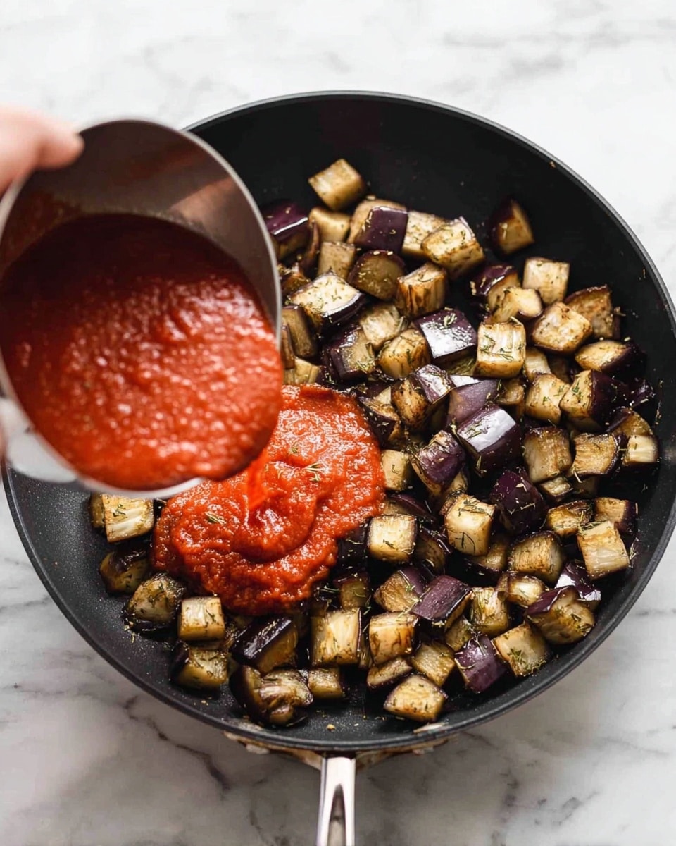 A black frying pan filled with small, evenly cut cubes of cooked eggplant that are a mix of dark purple skin and light beige flesh, lightly browned and seasoned with visible small herbs. A woman's hand is pouring thick, bright red tomato sauce from a silver measuring cup over the eggplants in the pan. The background is a white marbled surface. photo taken with an iphone --ar 4:5 --v 7